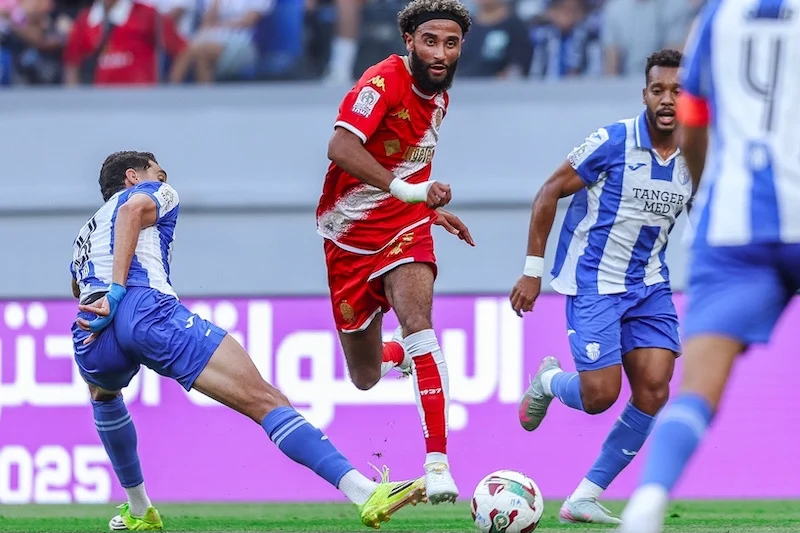 IRT-WAC1 | MCG24 | موقع إلكتروني مغربي شامل متجدد على مدار الساعة Soccer player in a red jersey leaps with the ball as blue-and-white defenders approach on a green pitch behind him, stadium spectators in the background.