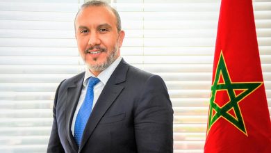 Professional portrait: man in a dark suit and blue tie beside a Moroccan flag by window blinds