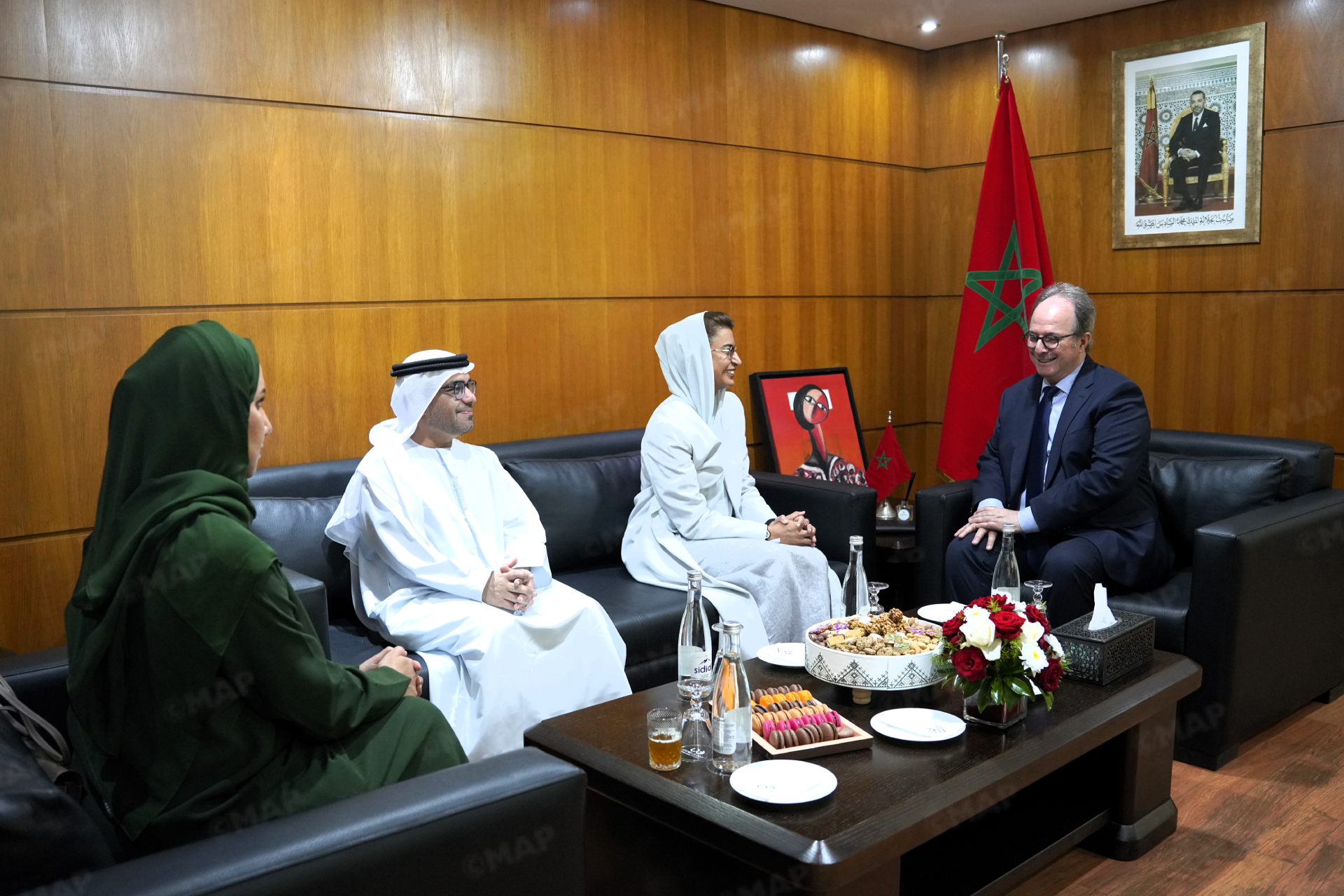 MAP6661 | MCG24 | موقع إلكتروني مغربي شامل متجدد على مدار الساعة Officials in traditional white robes and a woman in a light gray hijab sit on a leather sofa in a formal meeting room with a Moroccan flag nearby and a framed portrait on the wall.
