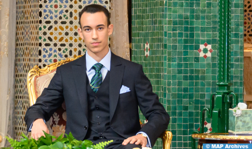 Moulay-El-Hassan1-1_jpg | MCG24 | موقع إلكتروني مغربي شامل متجدد على مدار الساعة Formal portrait of a man in a black suit and teal striped tie, seated on a gold chair beside a mosaic-tiled wall at an ornate setting.