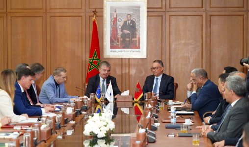Formal diplomatic meeting with officials seated around a long wooden table; Moroccan and Turkish flags on the table and a portrait hanging on a paneled wall behind.