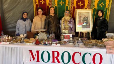 Nouveau-projet216 | MCG24 | موقع إلكتروني مغربي شامل متجدد على مدار الساعة Six women stand behind a Moroccan craft stall displaying metalware, pottery, textiles, and a framed portrait, with a colorful backdrop and a bold 'MOROCCO' banner.