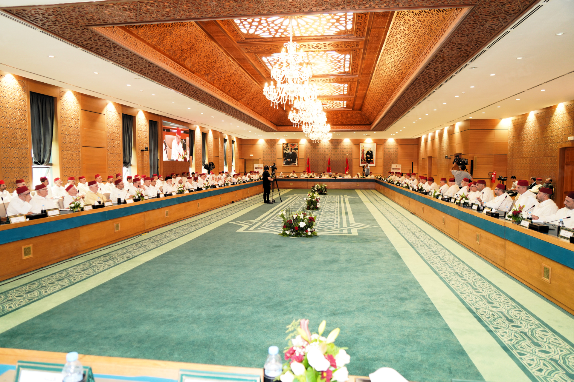 Wide-angle view of a formal conference in a grand room, delegates in white attire and red caps seated around a long U-shaped table under a crystal chandelier.