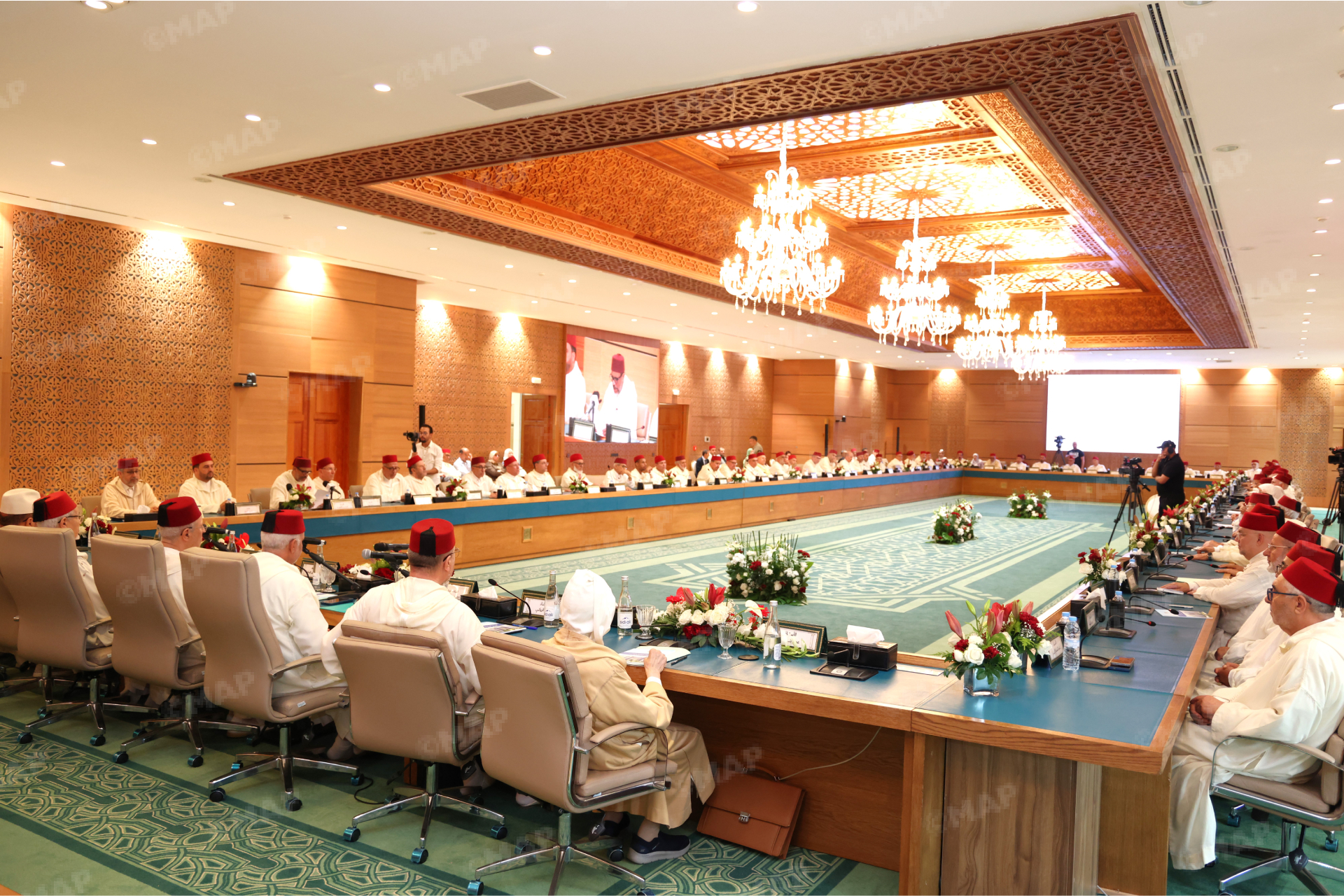 Formal meeting in a grand hall with delegates in white attire and red fez hats seated around a U-shaped table.