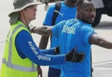 Pape-Thiaw-airport-usa | MCG24 | موقع إلكتروني مغربي شامل متجدد على مدار الساعة Airport worker in a neon vest guides a man wearing a blue Senegal shirt with arms extended on the tarmac.