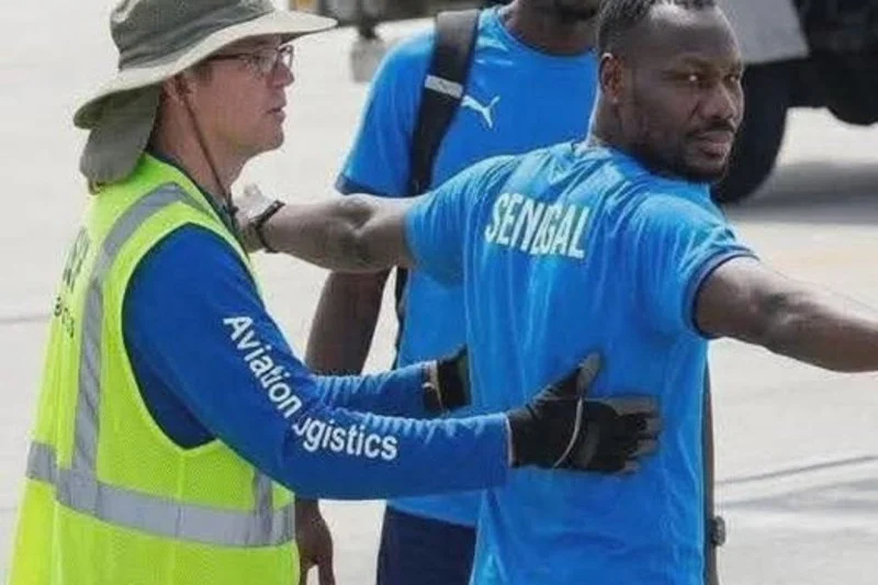 Pape-Thiaw-airport-usa | MCG24 | موقع إلكتروني مغربي شامل متجدد على مدار الساعة Airport worker in a neon vest guides a man wearing a blue Senegal shirt with arms extended on the tarmac.