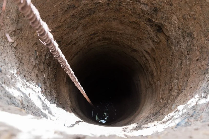 Puits-a-eau | MCG24 | موقع إلكتروني مغربي شامل متجدد على مدار الساعة Deep vertical well shaft with a rope stretched along the left side descending into darkness.