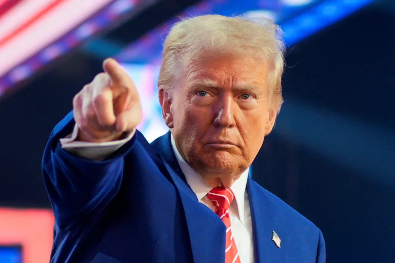 Man in a blue suit and red striped tie points toward the viewer on a stage, with a serious expression and bright lights in the background.