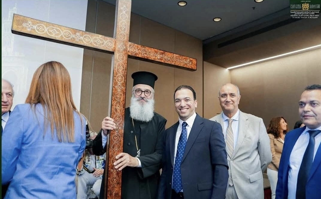 Group of formally dressed people posing indoors beside a large decorative wooden cross.