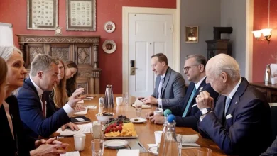 Staffan-de-Mistura-Massad-Boulos-Andreas-Motzfeldt-Kravik-1 | MCG24 | موقع إلكتروني مغربي شامل متجدد على مدار الساعة Group of professionals seated around a long wooden conference table in a formal red room, discussing documents with coffee and fruit platter in the center.