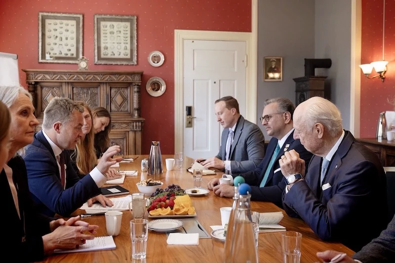 Staffan-de-Mistura-Massad-Boulos-Andreas-Motzfeldt-Kravik-1 | MCG24 | موقع إلكتروني مغربي شامل متجدد على مدار الساعة Group of professionals seated around a long wooden conference table in a formal red room, discussing documents with coffee and fruit platter in the center.