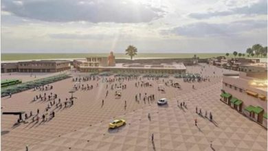 Wide view of a spacious coastal plaza with patterned tiles, people walking, a few cars, and low buildings along the edges.