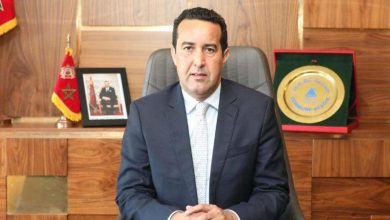WhatsApp-Image-2025-10-07-at-123923 | MCG24 | موقع إلكتروني مغربي شامل متجدد على مدار الساعة Man in a dark suit sits at a wooden desk in an office, facing the camera with hands folded on the desk.