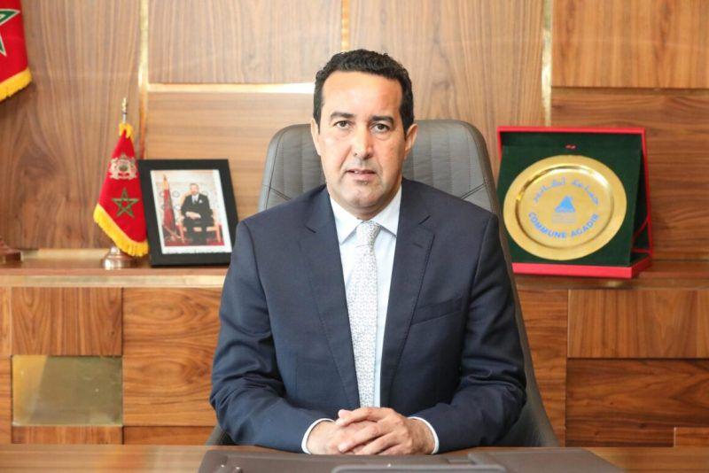 Man in a dark suit sits at a wooden desk in an office, facing the camera with hands folded on the desk.