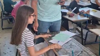 A girl with glasses sits at a desk writing on papers while a man stands nearby in a classroom filled with other students.