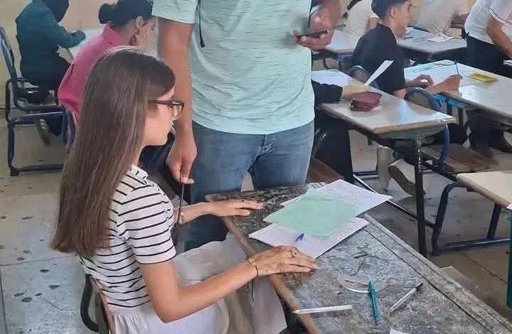 A girl with glasses sits at a desk writing on papers while a man stands nearby in a classroom filled with other students.