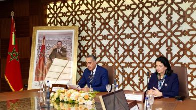 Two officials sit at a polished conference table in a room with a geometric wooden screen; a Moroccan flag and a large framed portrait are in the background.