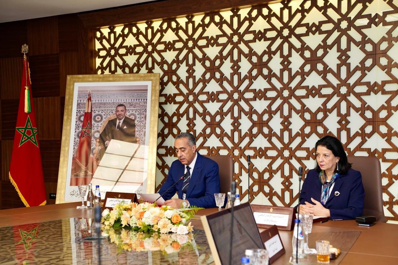 Two officials sit at a polished conference table in a room with a geometric wooden screen; a Moroccan flag and a large framed portrait are in the background.