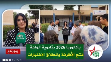 News reporter with a green microphone (MCG24) speaks to camera while a group gathers outside a campus building; green Arabic caption banner on screen with MCG24 logo in corner.