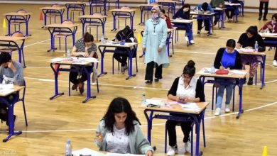 Exam session in a gym: students at single desks spaced apart, supervisor in a light blue coat wearing a mask.