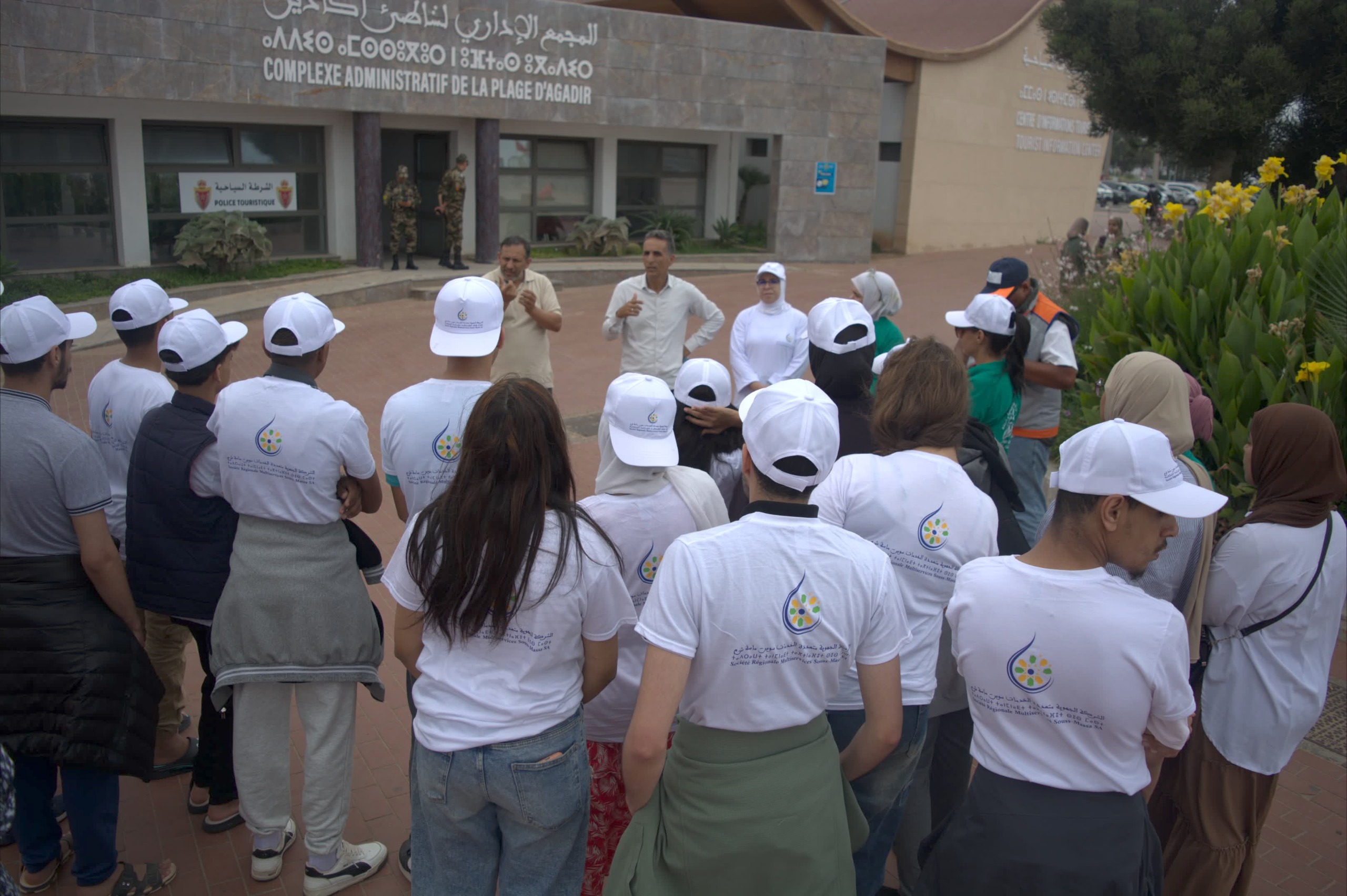 Group of people in white hats listening to two men speaking outside a police tourism office entrance.