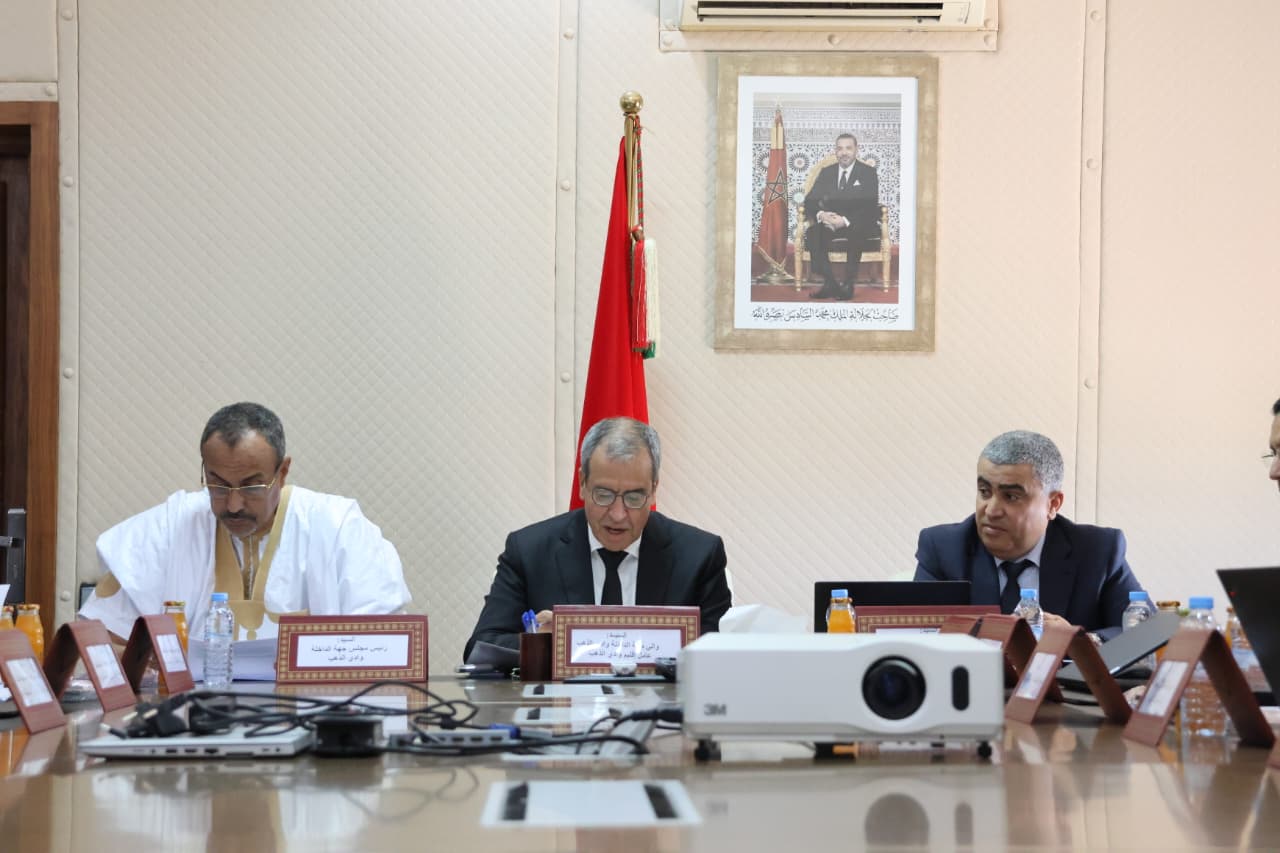 WhatsApp Image 2026-06-10 at 183449 | MCG24 | موقع إلكتروني مغربي شامل متجدد على مدار الساعة Three men in formal attire sit at a conference table with nameplates, a projector, and bottled drinks; Moroccan flag stands in the background near a framed portrait.