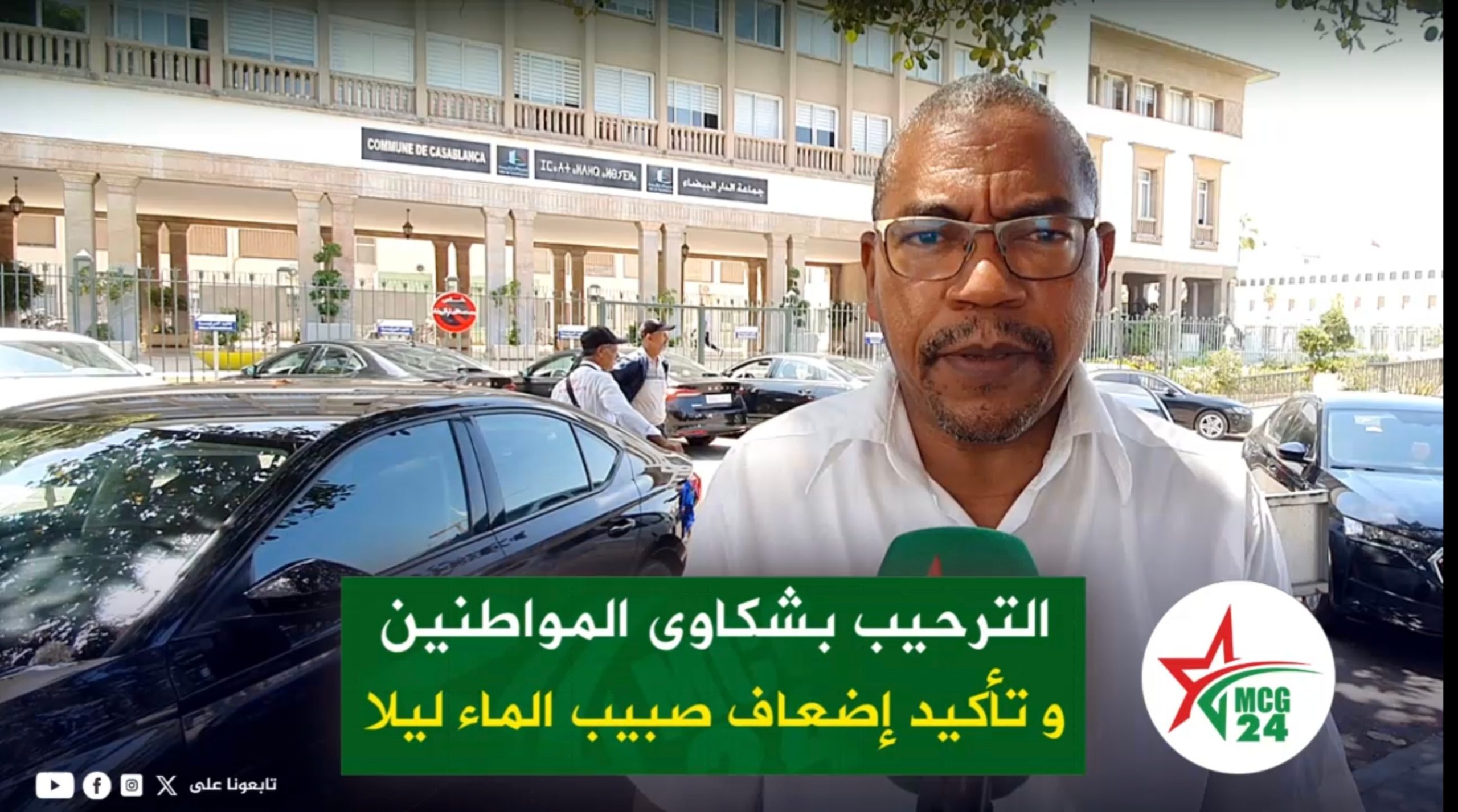 مفتاح: تخفيض صبيب المياه ليلا إجراء تقني.. و SRM البيضاء سطات منفتحة على شكايات المواطنين | MCG24 | موقع إلكتروني مغربي شامل متجدد على مدار الساعة Male reporter wearing glasses speaks to camera outside a government building, with a green Arabic banner at the bottom and parked cars behind him.