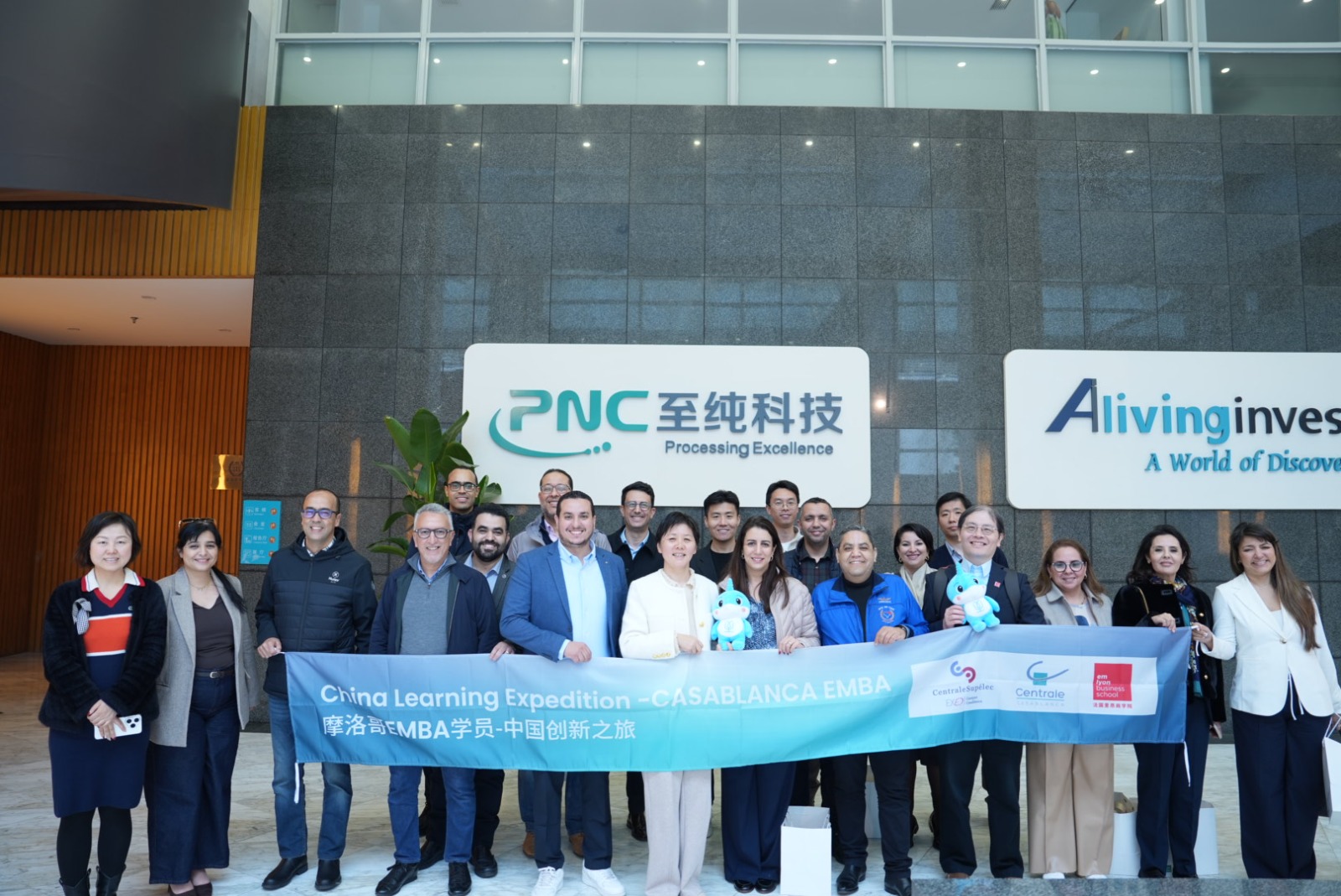 Group of diverse professionals posing for a photo in a modern lobby, holding a blue banner that reads 'China Learning Expedition – Casablanca EMBA' with PNC and AlivingInvest signs in the background.