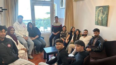 A diverse group of young adults seated and standing in a living room, posing for a group photo near a window and sofa.