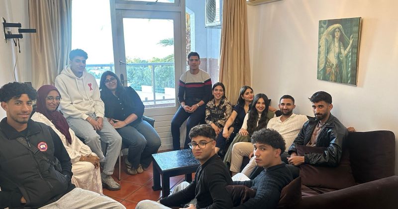 WhatsApp Image 2026-06-12 at 185359 | MCG24 | موقع إلكتروني مغربي شامل متجدد على مدار الساعة A diverse group of young adults seated and standing in a living room, posing for a group photo near a window and sofa.