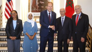 Five officials in formal attire pose on a marble staircase, with US and Moroccan flags and a framed portrait behind them; central man and partner shake hands.