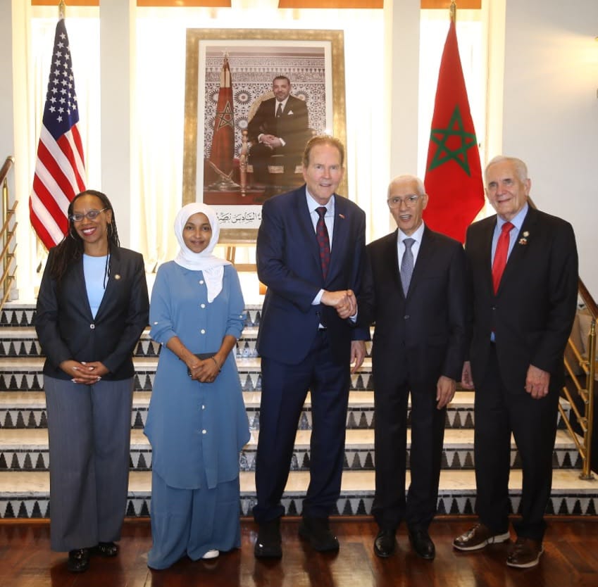 Five officials in formal attire pose on a marble staircase, with US and Moroccan flags and a framed portrait behind them; central man and partner shake hands.