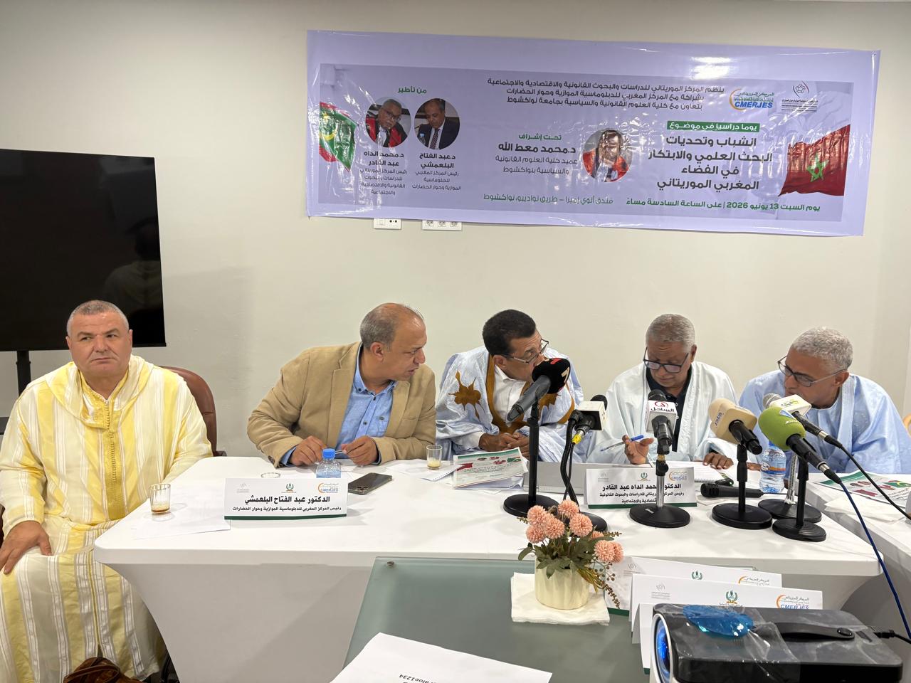 Panel of five men seated at a conference table with multiple microphones, a banner with Arabic text behind them, and name placards in front of the panelists.