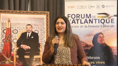 Woman speaker at a podium delivering a talk, with a large banner reading 'Forum de l'Atlantique' in the background and a framed portrait of a man beside a Moroccan flag to her left.