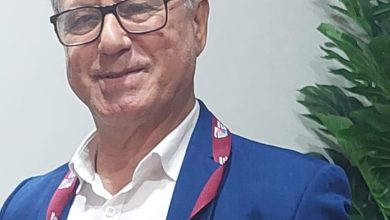 WhatsApp Image 2026-06-16 at 184734 | MCG24 | موقع إلكتروني مغربي شامل متجدد على مدار الساعة Smiling man in a blue suit and white shirt wearing glasses, with a maroon lanyard and badge, indoors near a large green plant.