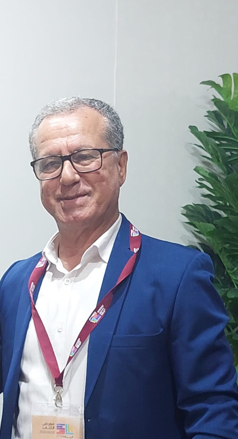 WhatsApp Image 2026-06-16 at 184734 | MCG24 | موقع إلكتروني مغربي شامل متجدد على مدار الساعة Smiling man in a blue suit and white shirt wearing glasses, with a maroon lanyard and badge, indoors near a large green plant.
