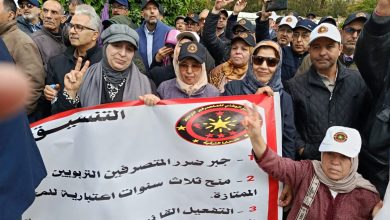 Crowd of protesters holding a banner with Arabic text and a circular emblem; several people raise their hands, some making peace signs.