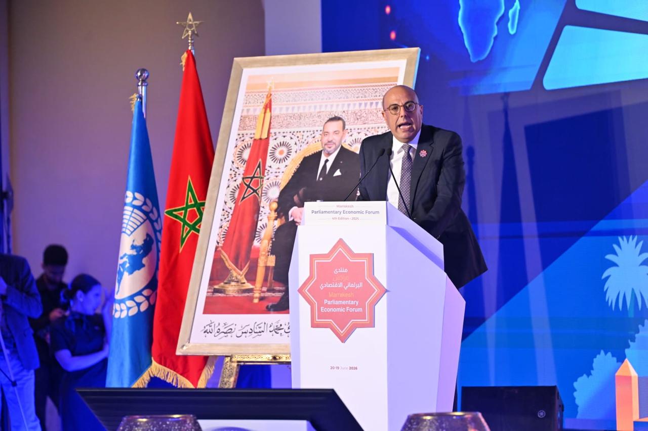 Speaker at a podium delivering a speech at a Parliamentary Economic Forum, with the Moroccan flag and a blue UN-style flag beside him, and a large framed portrait in the background.