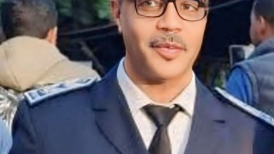 Young man in a formal police uniform with a blue cap, glasses, and tie, outdoors.