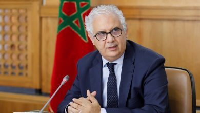 WhatsApp Image 2026-06-19 at 132921 | MCG24 | موقع إلكتروني مغربي شامل متجدد على مدار الساعة Middle-aged man with gray hair and glasses in a navy suit speaks into a microphone at a formal meeting, Moroccan flag in the background.