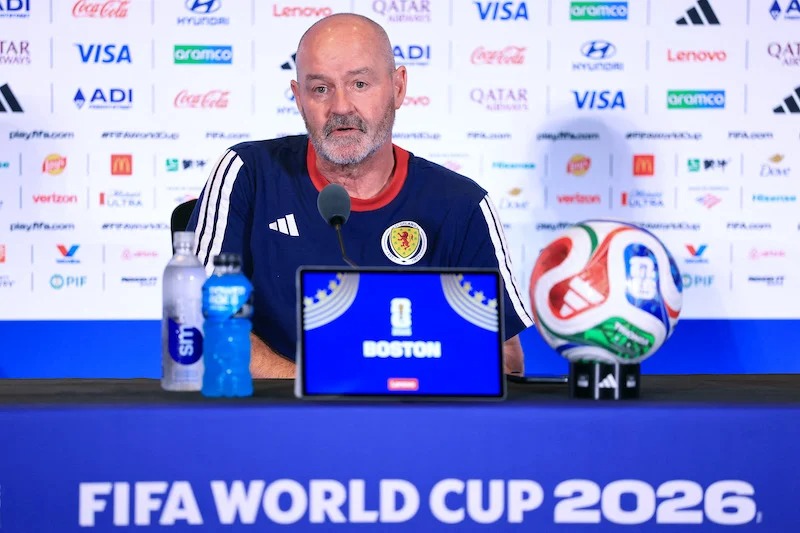 Male sports official at a World Cup press conference; the table displays a blue sign reading 'BOSTON', with a soccer ball on the right and a World Cup 2026 backdrop behind him.