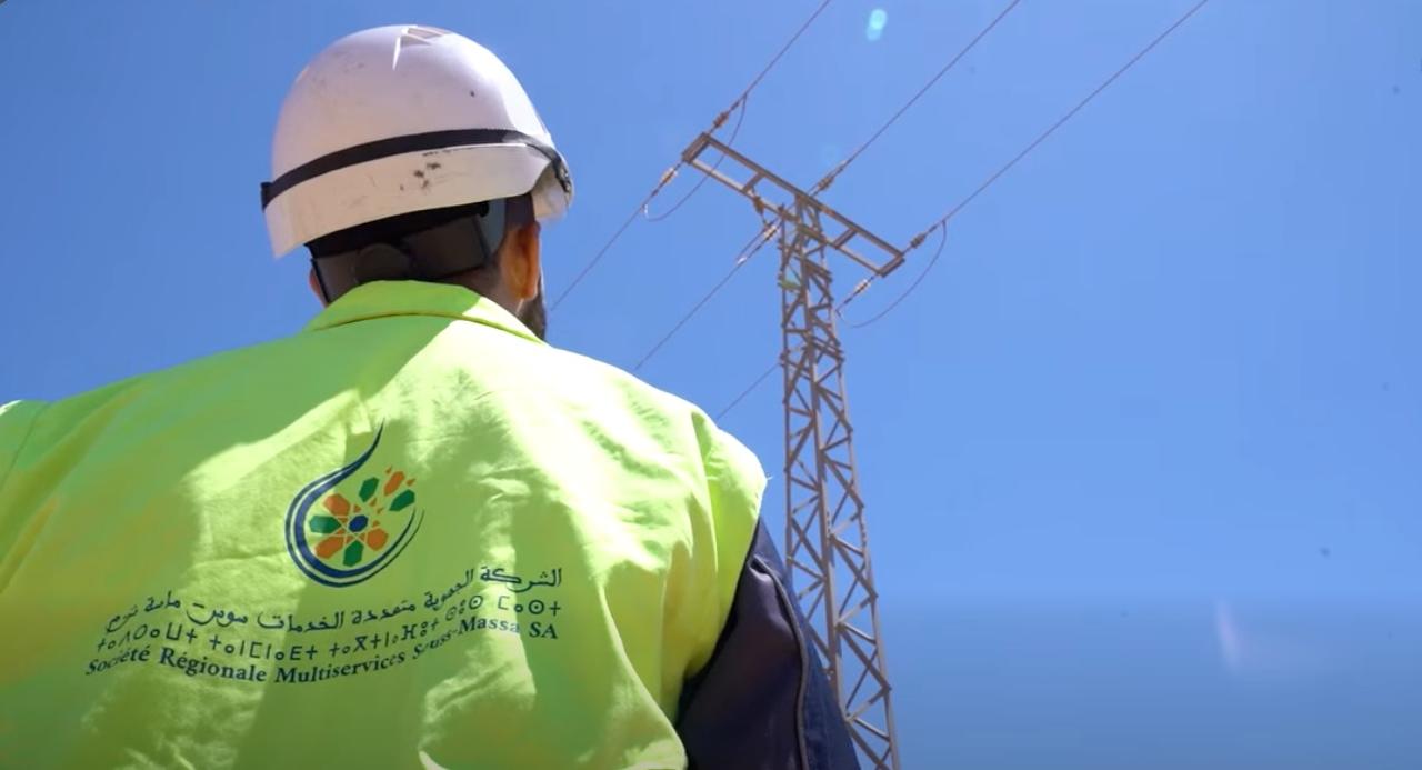WhatsApp Image 2026-06-21 at 180450 | MCG24 | موقع إلكتروني مغربي شامل متجدد على مدار الساعة Back view of a worker in a neon safety jacket and white helmet inspecting a high-voltage power line tower against a blue sky.