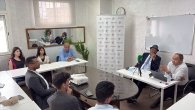 Panel discussion in a bright meeting room with two men at a table with microphones and a laptop, while several attendees listen at white tables and a projector sits on a round table in the foreground.
