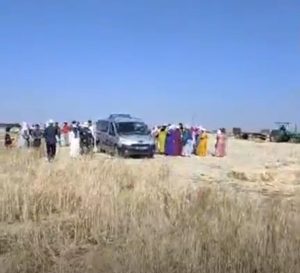 Group of people in colorful robes gathered around a silver van in a dry, open field under a clear blue sky. Nearby vehicles are visible in the distance.