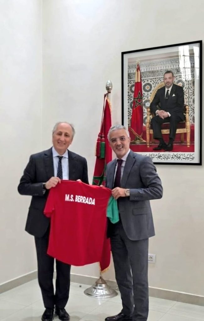 WhatsApp Image 2026-06-21 at 220231 | MCG24 | موقع إلكتروني مغربي شامل متجدد على مدار الساعة Two men in suits pose while holding a red jersey labeled'M.S. BERRADA' in a formal room with a Moroccan flag and a framed portrait of the Moroccan king on the wall.