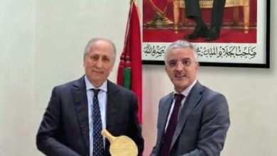 WhatsApp Image 2026-06-21 at 220232 1 | MCG24 | موقع إلكتروني مغربي شامل متجدد على مدار الساعة Two men in suits hold a golden curved trophy together in a formal room, with a Moroccan flag and a large portrait on the wall behind them.
