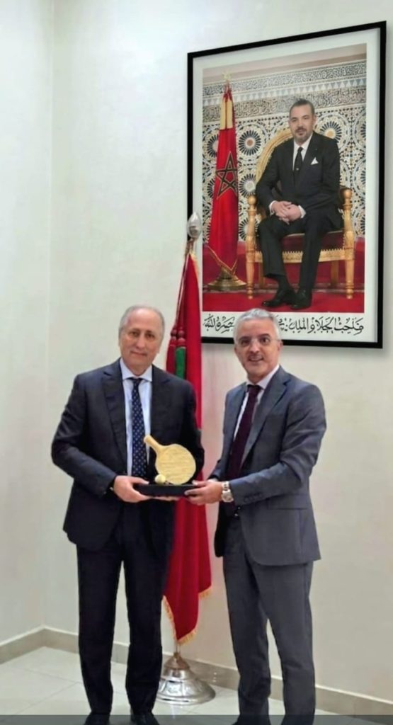 WhatsApp Image 2026-06-21 at 220232 1 | MCG24 | موقع إلكتروني مغربي شامل متجدد على مدار الساعة Two men in suits hold a golden curved trophy together in a formal room, with a Moroccan flag and a large portrait on the wall behind them.