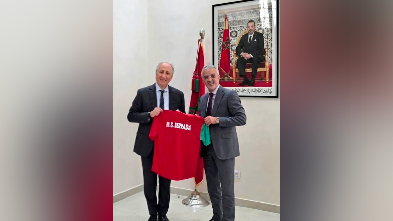 Two men in suits hold up a red jersey reading 'M.S. BERRADA' in a formal room with a Moroccan flag and a framed portrait in the background.