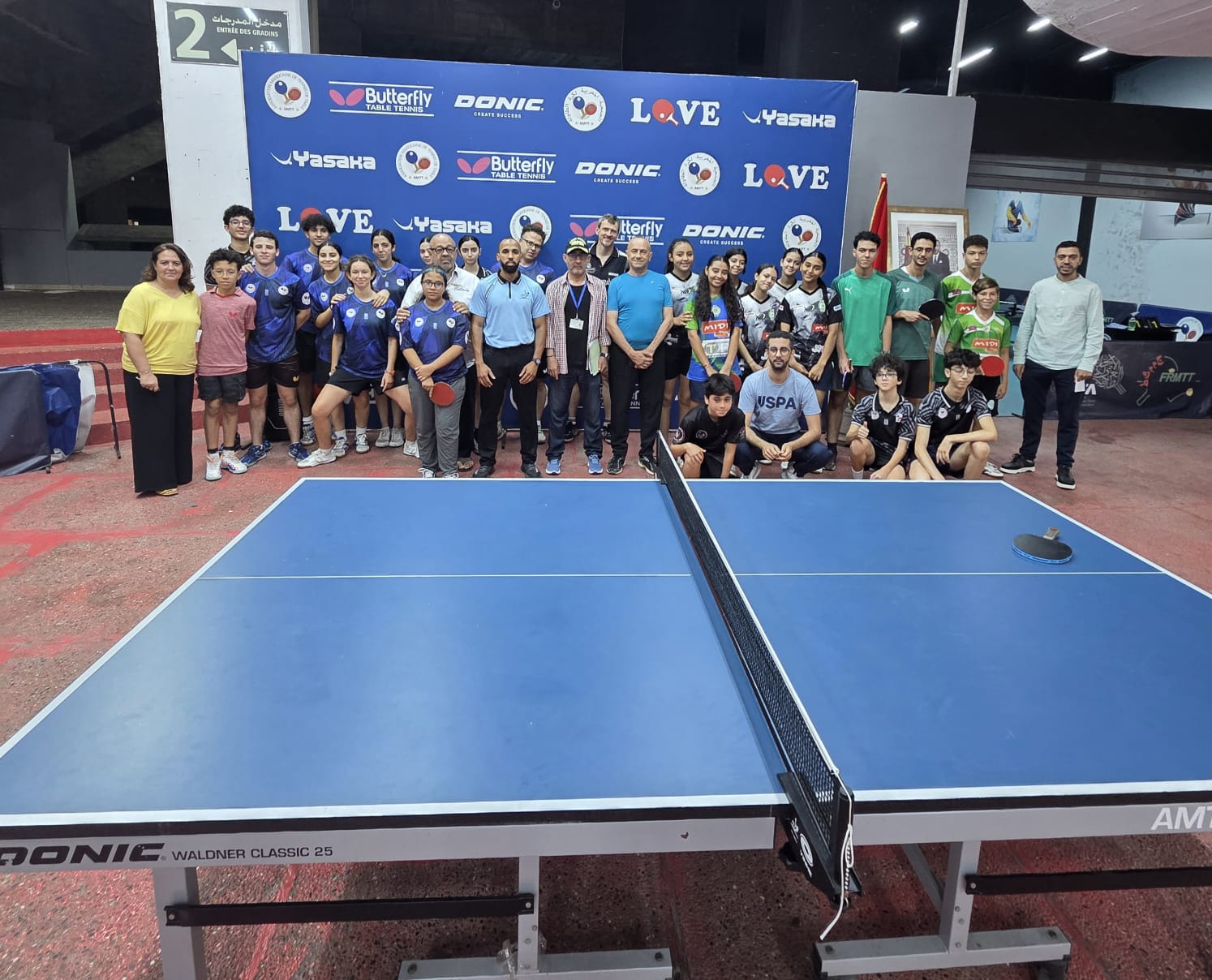 WhatsApp Image 2026-06-22 at 001436 | MCG24 | موقع إلكتروني مغربي شامل متجدد على مدار الساعة Group of young table tennis players and coaches posing for a photo behind a blue table tennis table inside a sports hall, with a branded backdrop featuring logos.