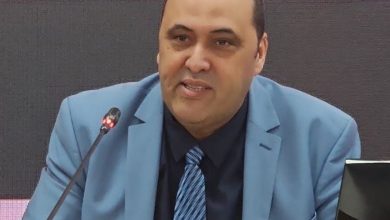 WhatsApp Image 2026-06-22 at 120859 | MCG24 | موقع إلكتروني مغربي شامل متجدد على مدار الساعة Man in a light-blue suit and striped tie speaks into a microphone at a conference podium, gesturing with his hand.